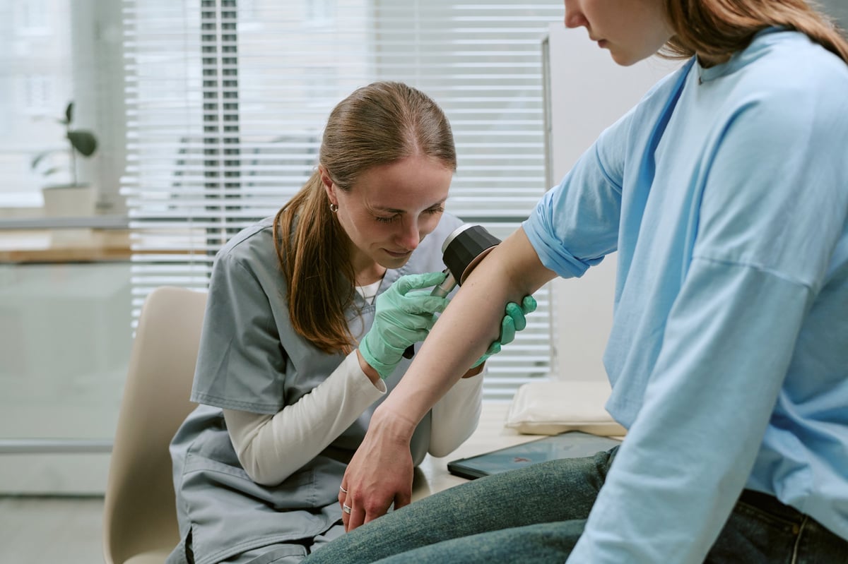 Young adult clinician examining woman arm with handheld diagnostic device during dermatology checkup